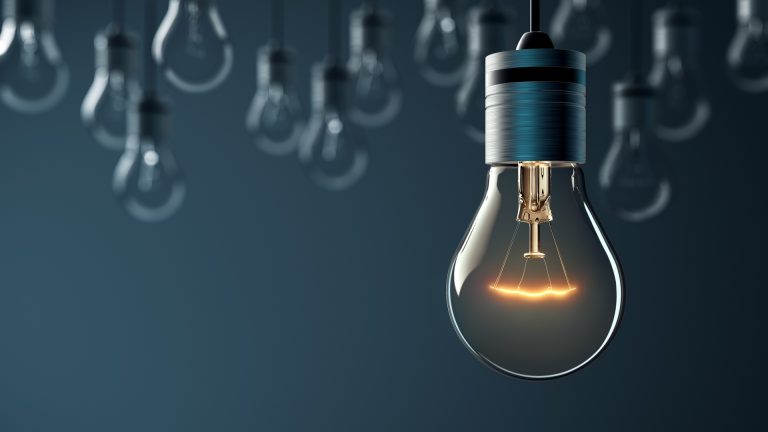 Image: On a deep blue background, light bulbs hang down on black coloured chords, with one in the foreground lit up shining bright white